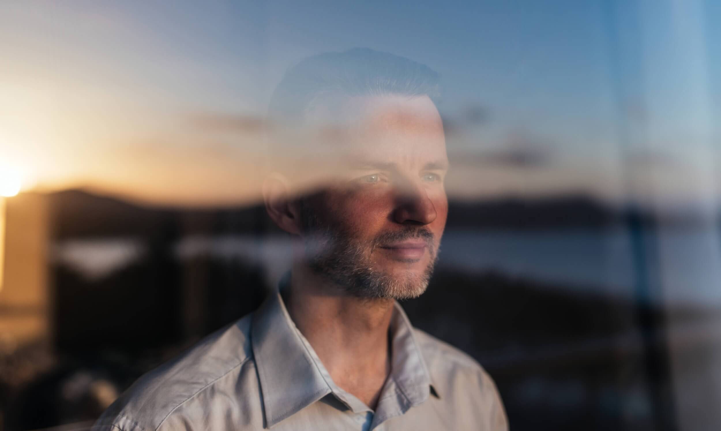 Photograph of man in white collared shirt looking out a window. Sunlight is reflected back onto him. 