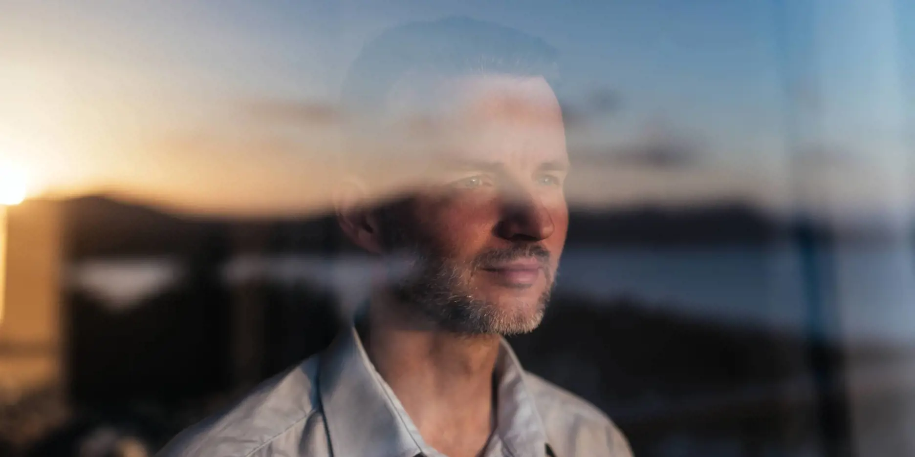 Photograph of man in white collared shirt looking out a window. Sunlight is reflected back onto him.