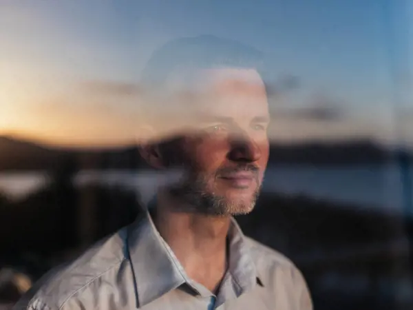 Photograph of man in white collared shirt looking out a window. Sunlight is reflected back onto him.