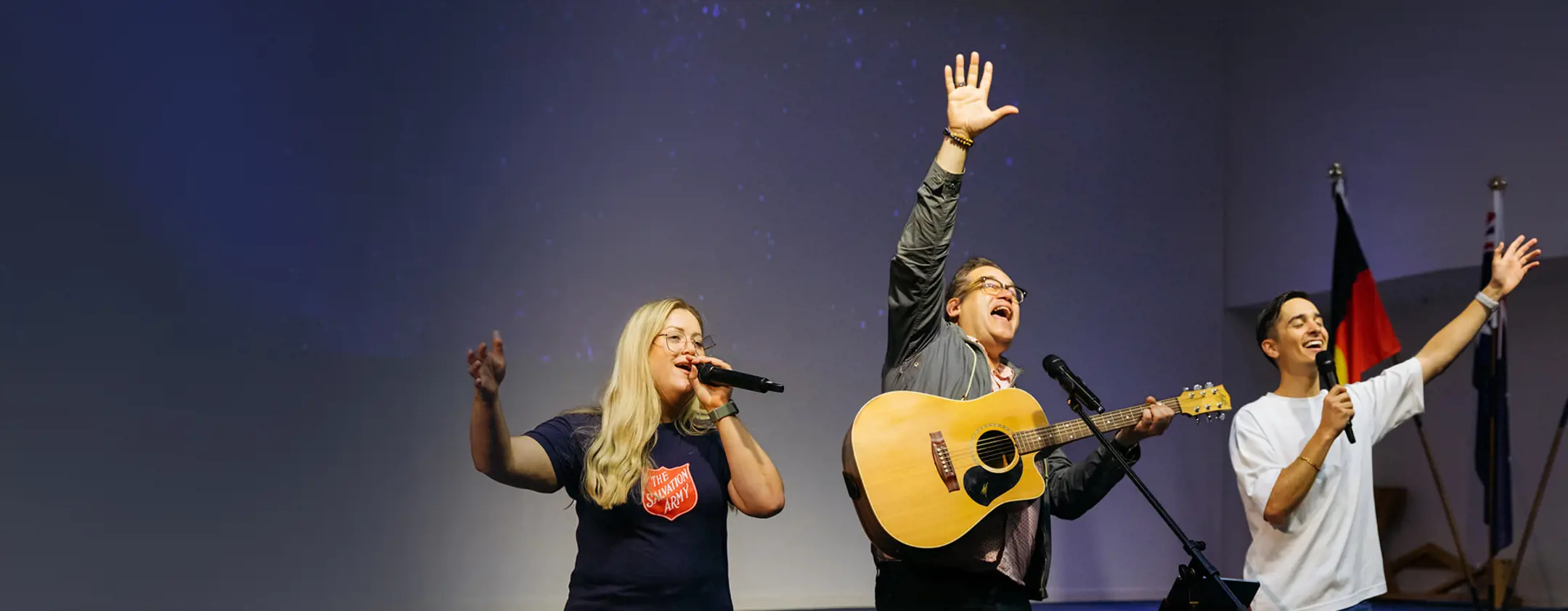 Three Salvos members on stage, woman in the Salvos shirt is singing into the microphone, the man in the middle is playing guitar with hand raised and the man on the right is singing with arm outstretched.