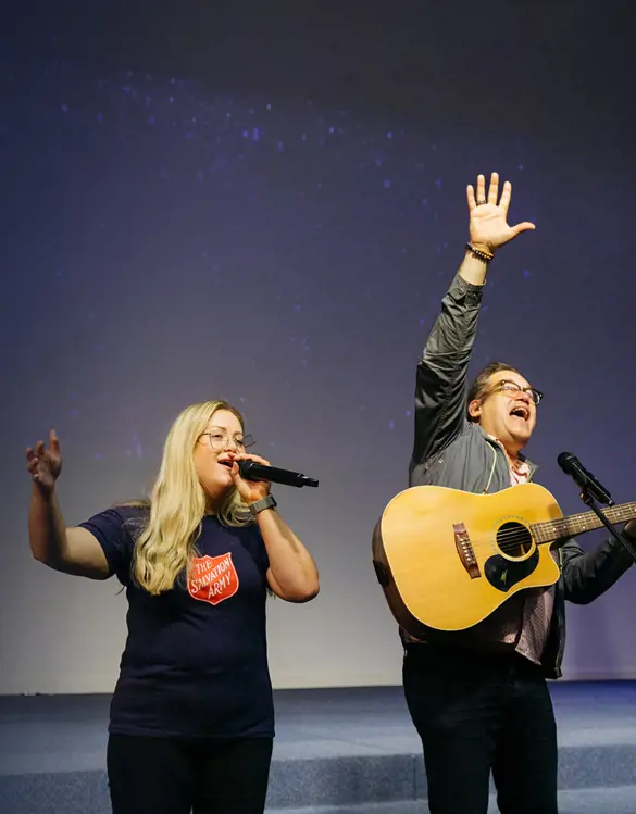 Three Salvos members on stage, woman in the Salvos shirt is singing into the microphone, the man in the middle is playing guitar with hand raised and the man on the right is singing with arm outstretched.
