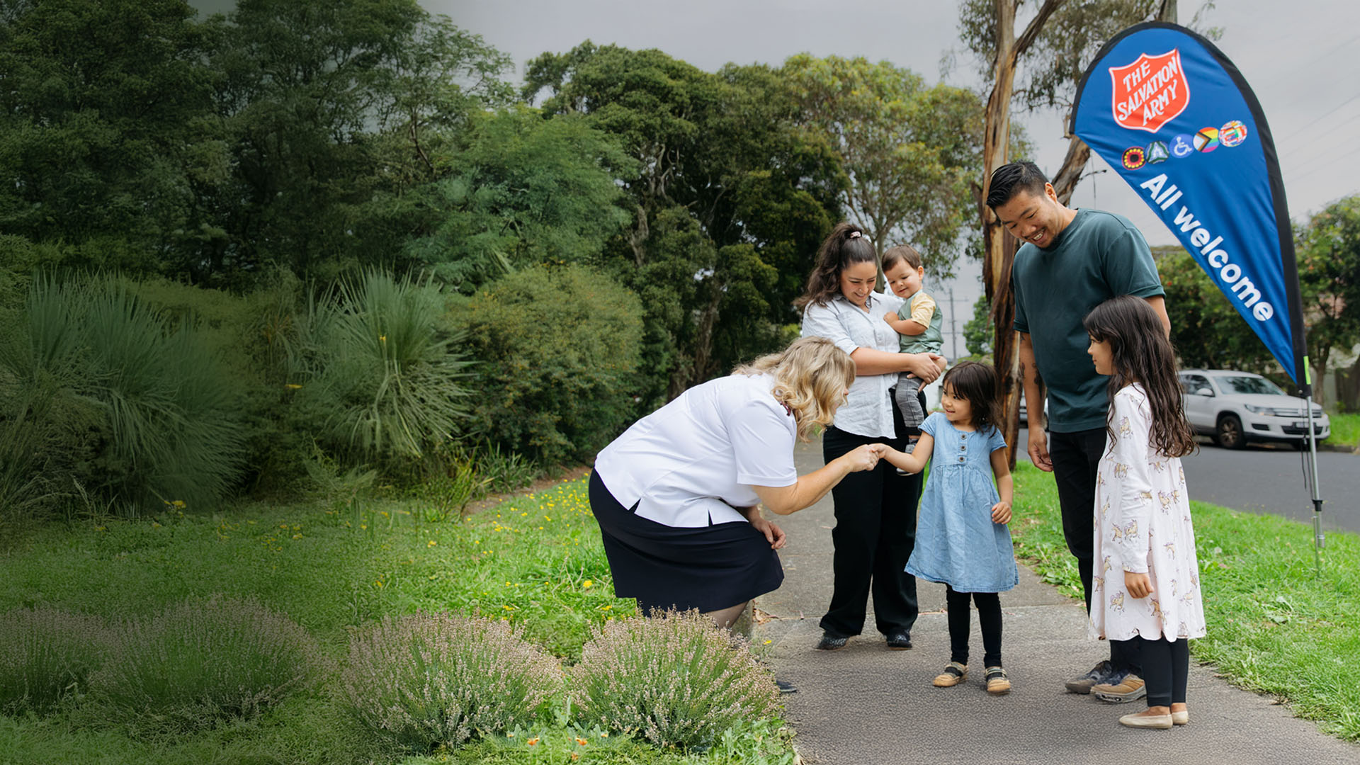 Female Salvos officer shaking hands with a girl and her family. The background is a path with surrounding grass and plants with a Salvos flag that says "All welcome".