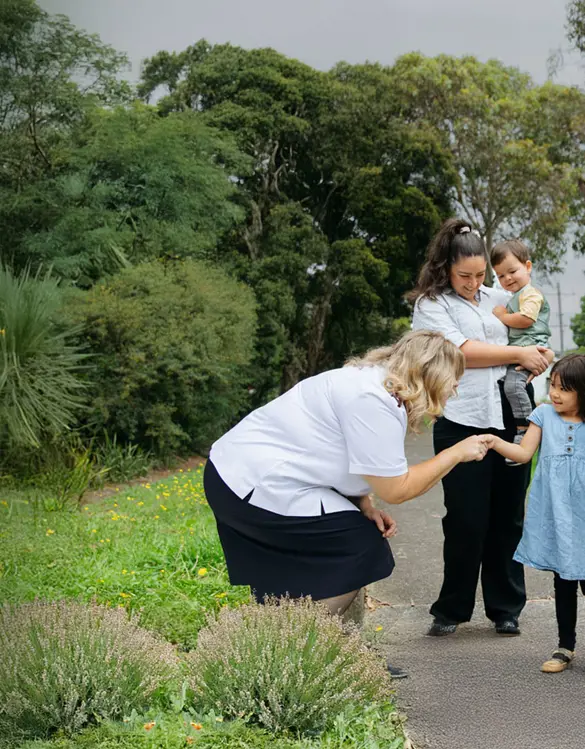 Female Salvos officer shaking hands with a girl and her family. The background is a path with surrounding grass and plants with a Salvos flag that says "All welcome".