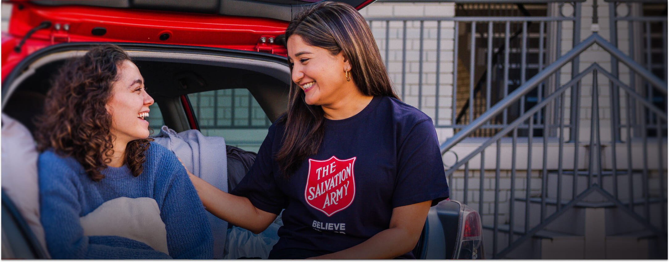 Female Salvos Officer supporting a young woman sitting in the boot of her car.