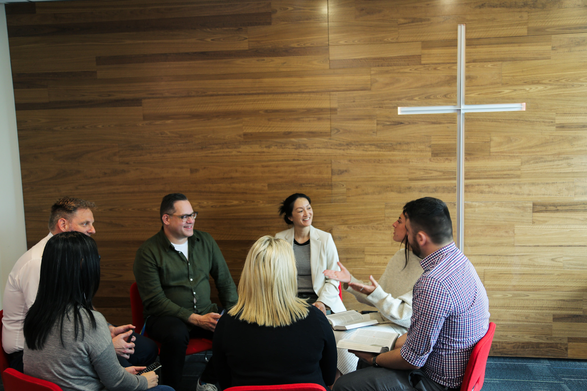 Six people sitting on red chairs in circle in active discussion with each other. Behind them is a white Christian Cross.