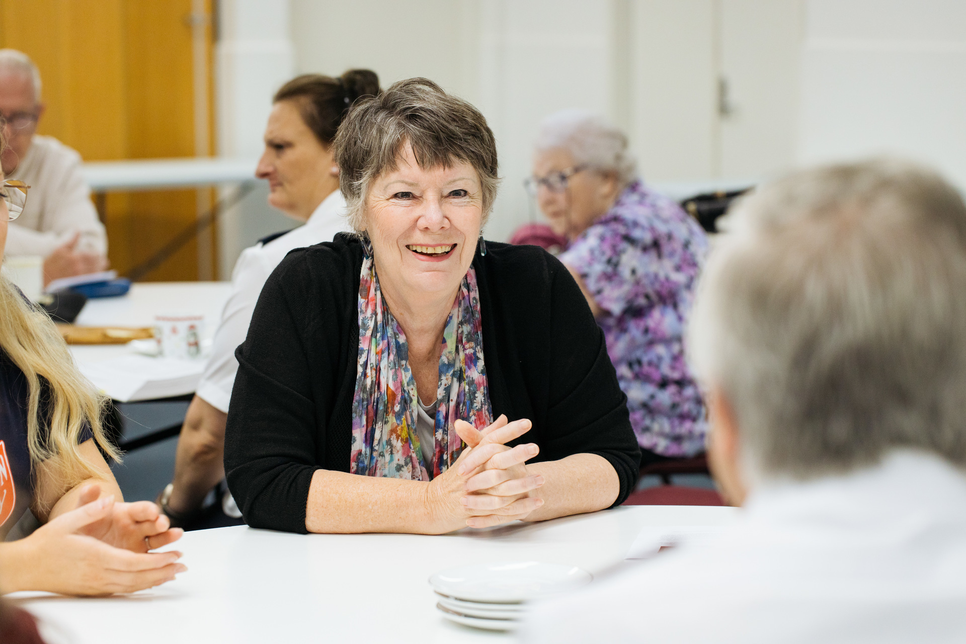 People sit around a table in a communal space. The focus is on a elderly woman who is smiling.