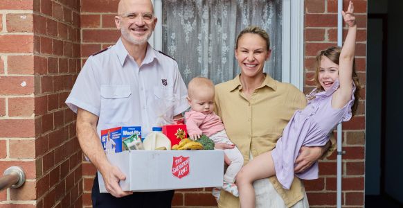 Amy smiling with her two daughters in her arms, standing next to a Salvos personnel carrying a hamper filled with groceries.