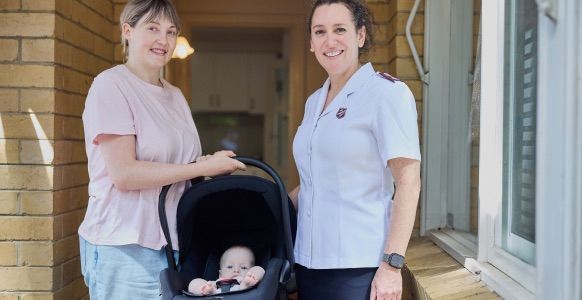 Emily standing with her young baby in a pram, smiling, next to a Salvos personnel