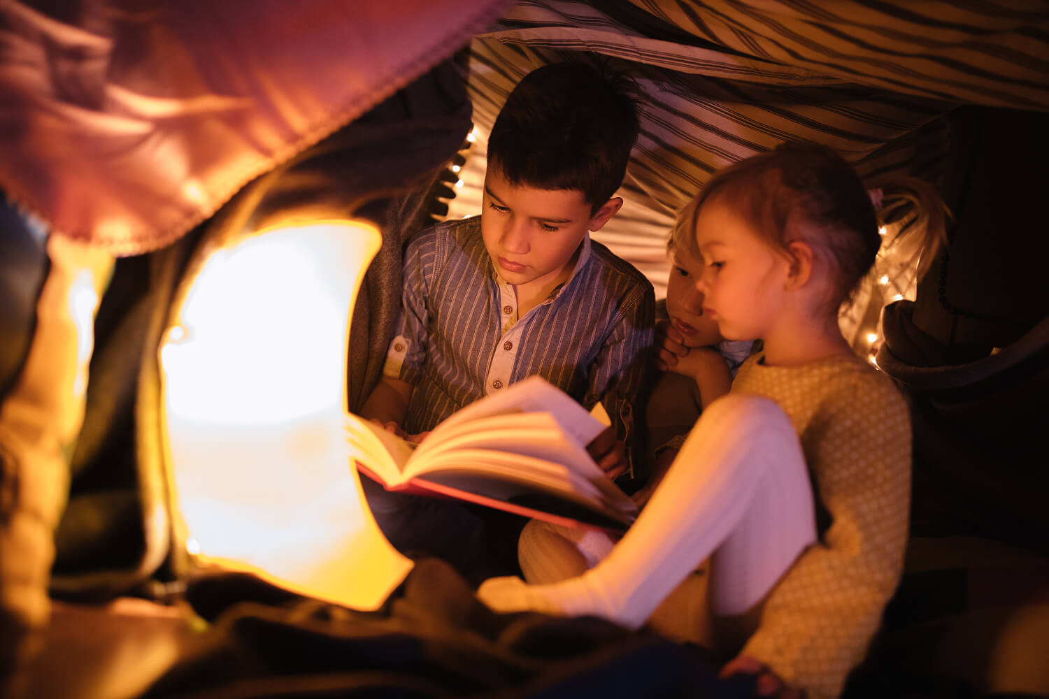 Three young children reading a book together inside a cozy blanket fort, softly illuminated by a warm night light.