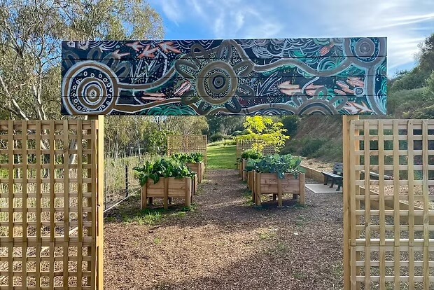 Community garden entrance with Aboriginal mural sign, raised garden beds, and surrounding bushland.