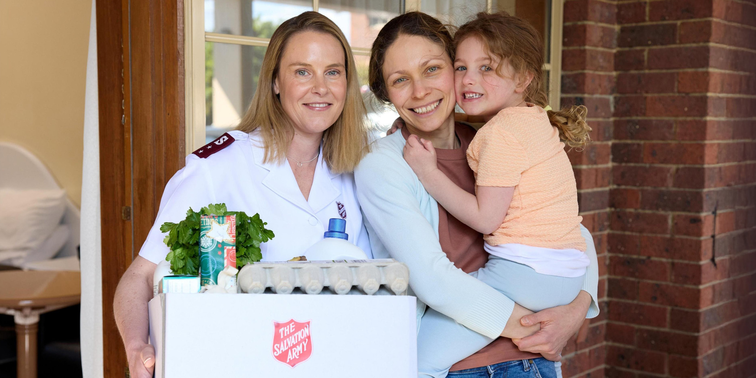 Salvos personnel holding a Christmas hamper standing next to a mother and daughter, smiling.