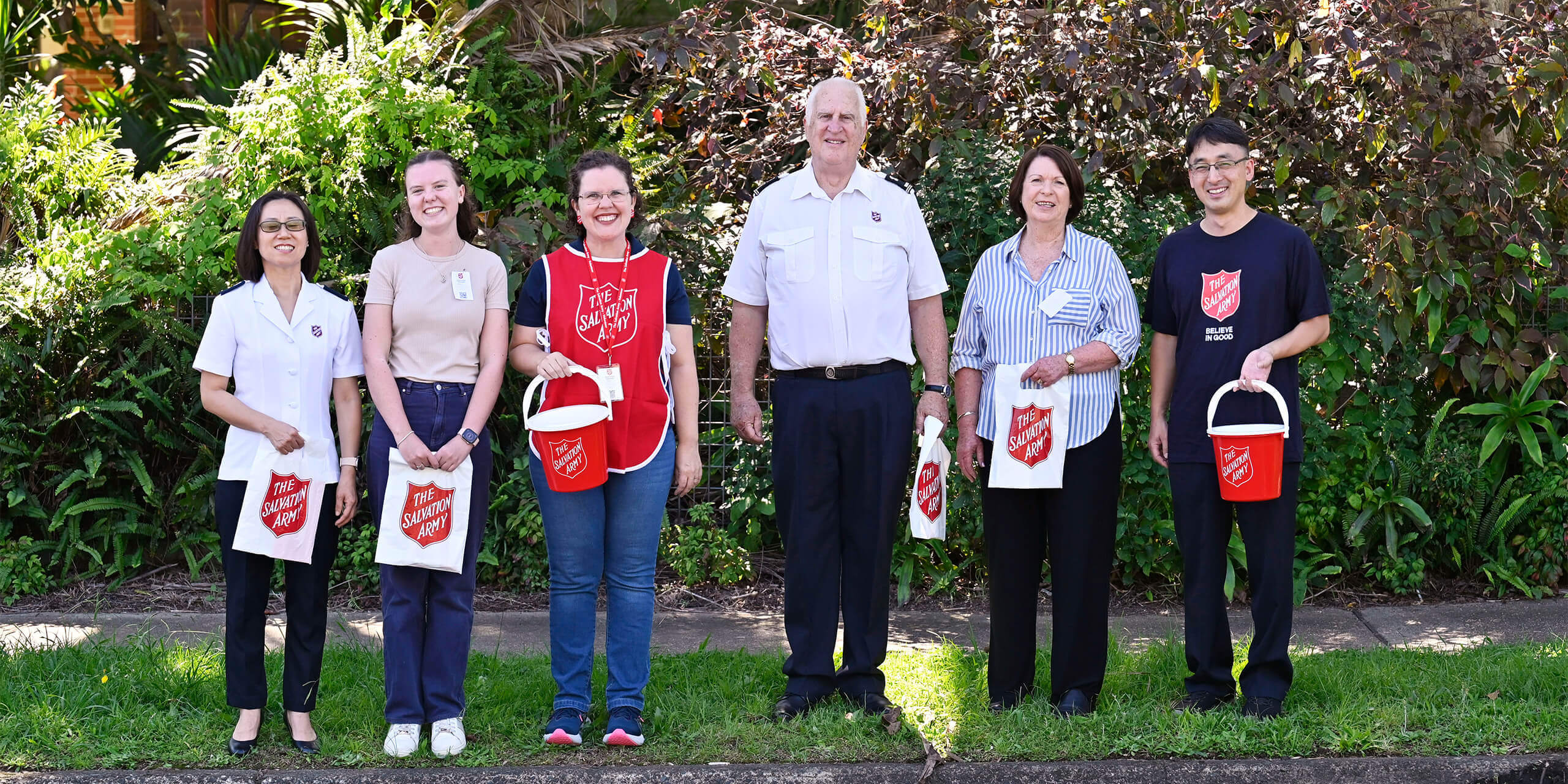 Red Shield Appeal volunteers smiling and holding a red bucket, standing next to a Salvos personnel.