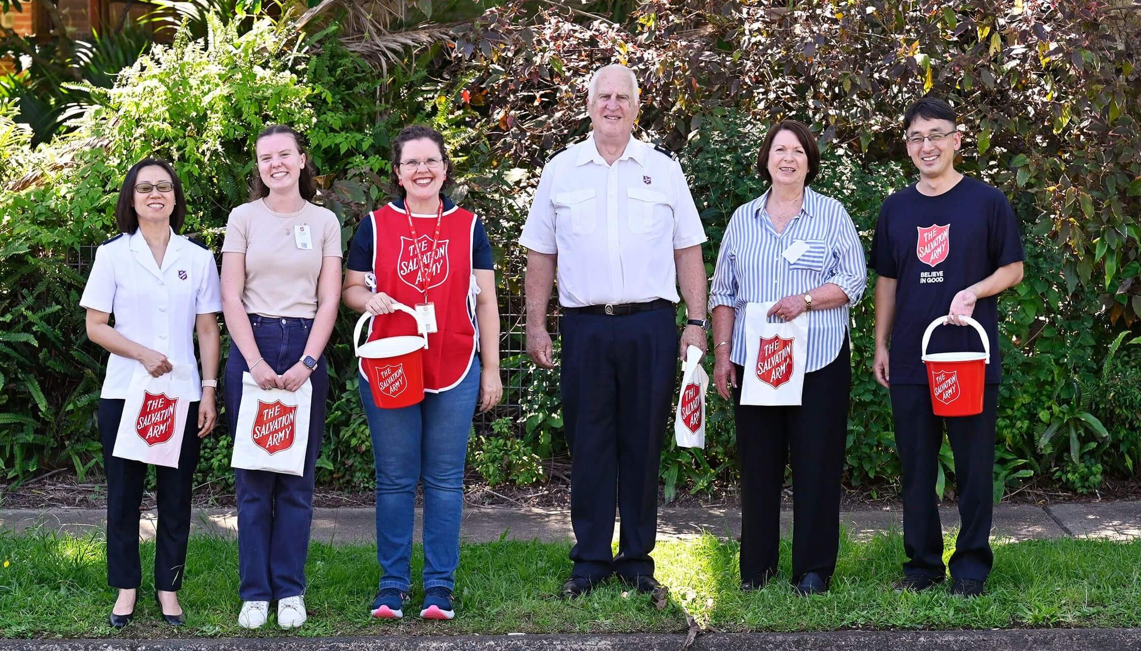 Red Shield Appeal volunteers smiling and holding a red bucket, standing next to a Salvos personnel.