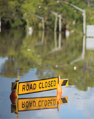 A flood scene. A 'road closed' sign is almost submerged by flood waters in what was once a street.