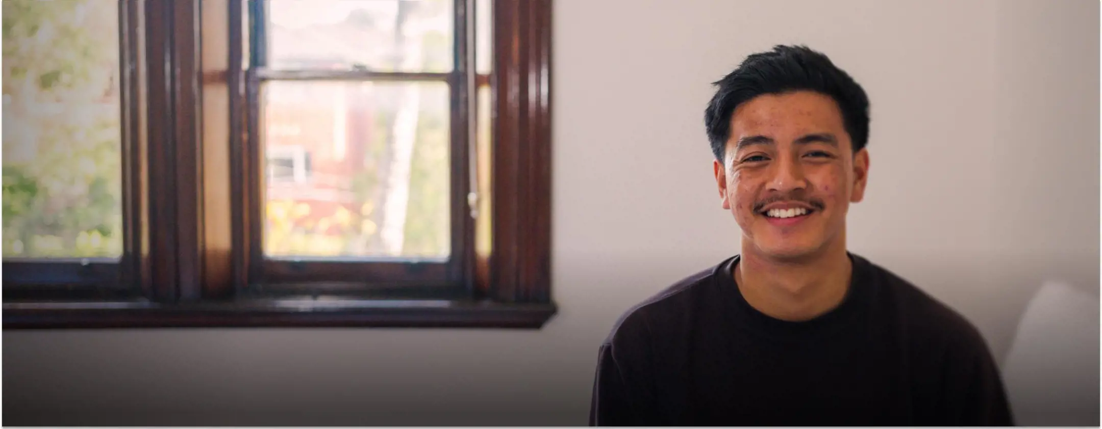 A young man smiles, sitting on a bed in a crisp clean room.