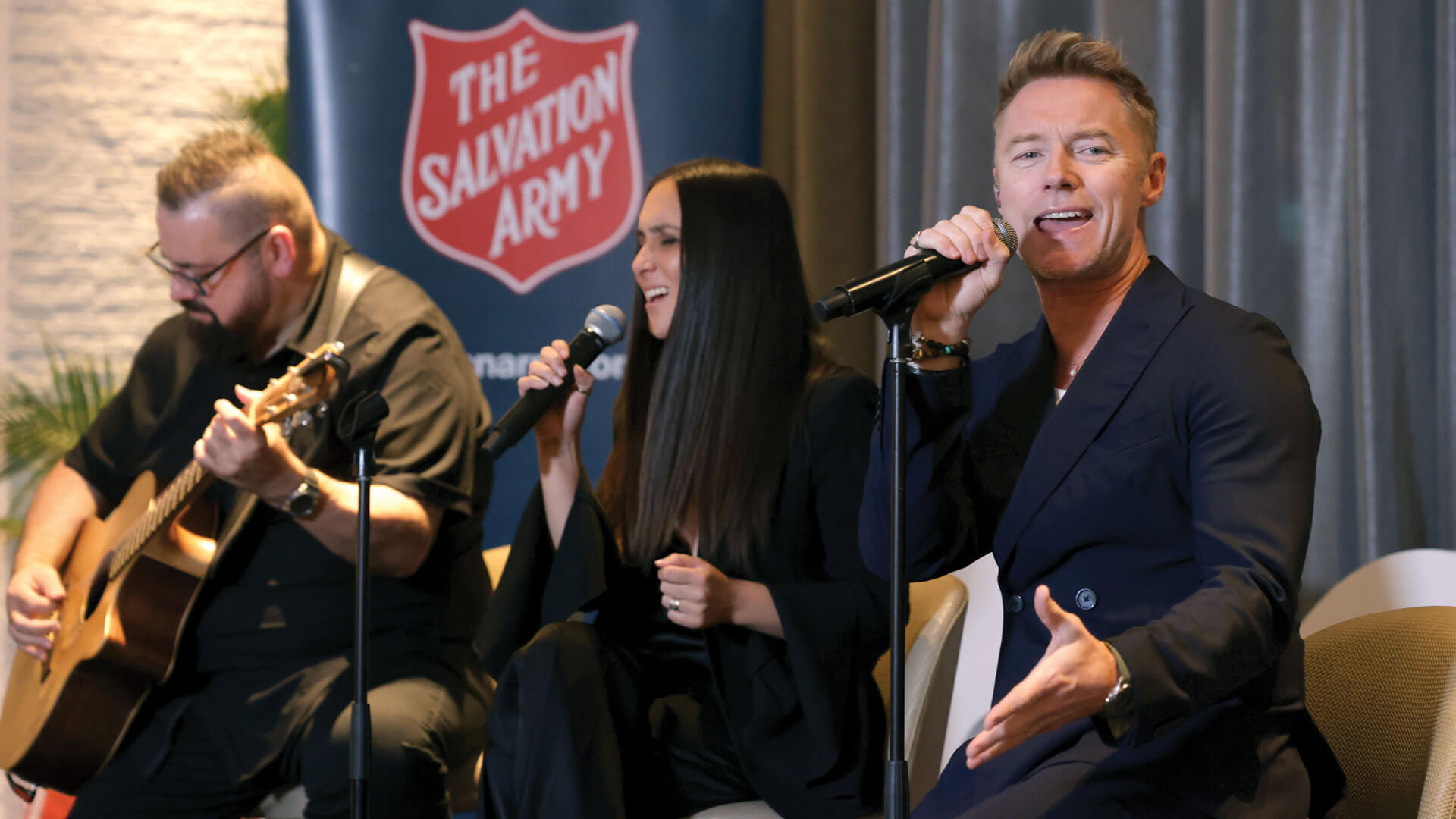 Ronan Keating is crooning with the help of another singer and a guitarist with a Salvos shield in the background.
