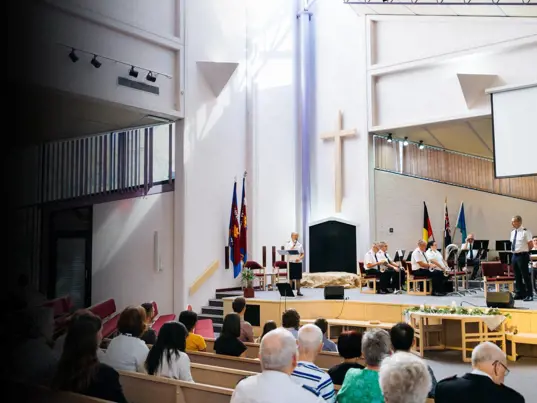 A wide view a brightly lit church service. An officers speaks from the stage to the congregation in our foreground.