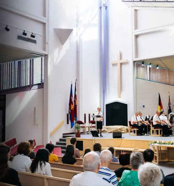A wide view a brightly lit church service. An officers speaks from the stage to the congregation in our foreground.