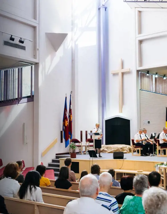 A wide view a brightly lit church service. An officers speaks from the stage to the congregation in our foreground.