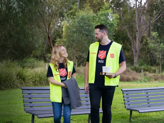 Two Salvation Army volunteers sharing a friendly conversation in a park, ready with warm coffee, blankets, and open hearts to support those doing it tough.