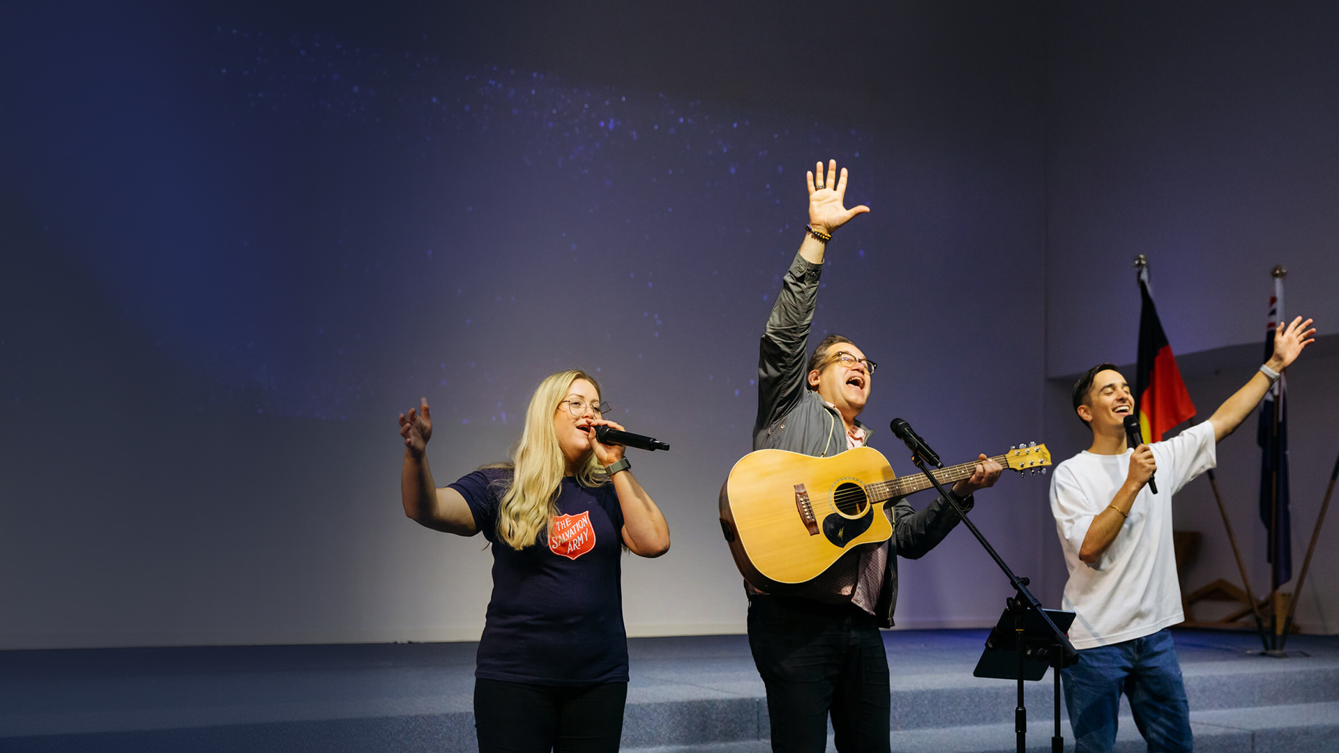 Three people leading worship on a church stage — one playing acoustic guitar, and the other 2 singing in a Salvation Army t-shirt, as heartfelt music fills the space.