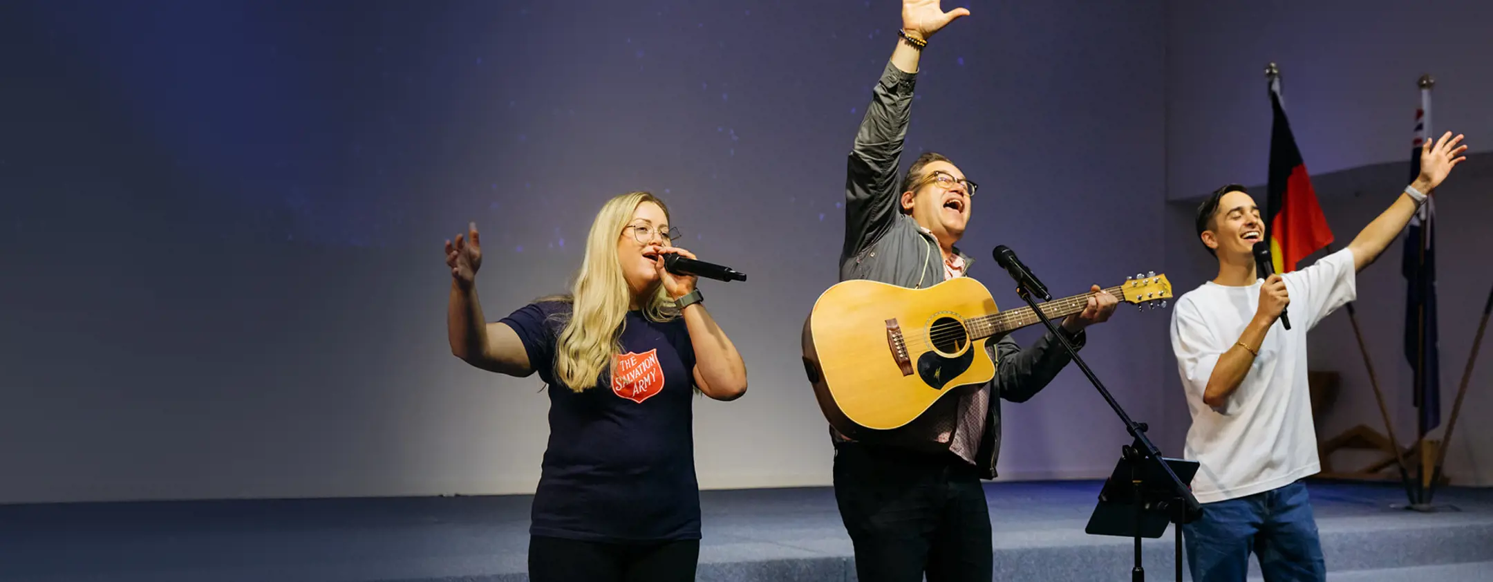 Three people leading worship on a church stage — one playing acoustic guitar, and the other 2 singing in a Salvation Army t-shirt, as heartfelt music fills the space.