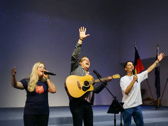 Three people leading worship on a church stage — one playing acoustic guitar, and the other 2 singing in a Salvation Army t-shirt, as heartfelt music fills the space.