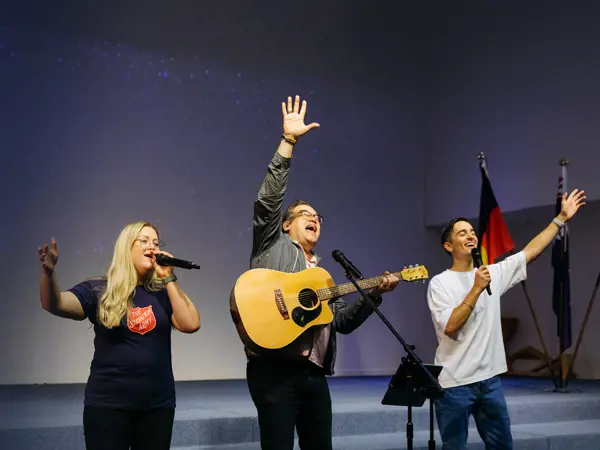 Three people leading worship on a church stage — one playing acoustic guitar, and the other 2 singing in a Salvation Army t-shirt, as heartfelt music fills the space.