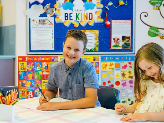 Two children sitting at a table smiling while drawing on paper.