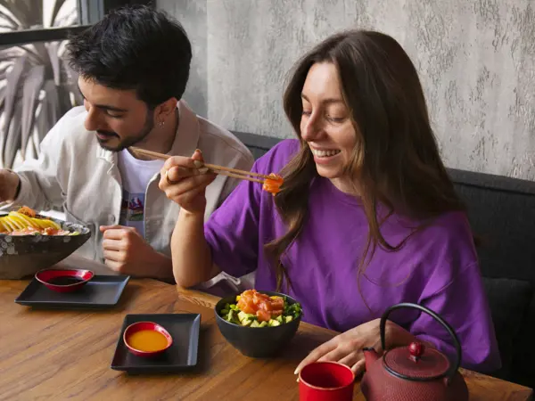 Two happy people eating salmon dishes with chopsticks at a restaurant table.