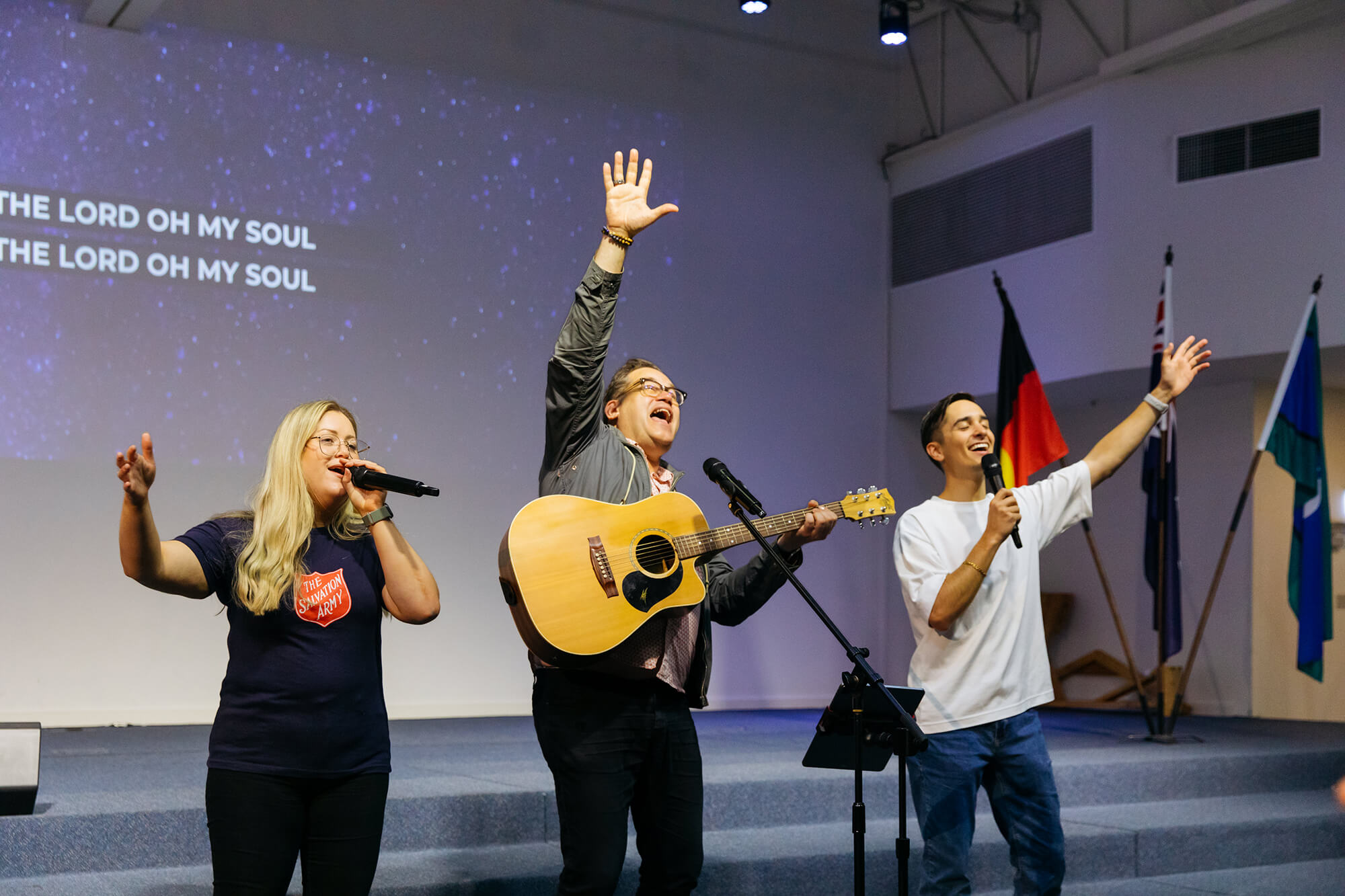 Three people sing with spirit, accompanied by one person playing the guitar.