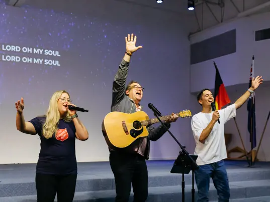 Three people sing with spirit, accompanied by one person playing the guitar.