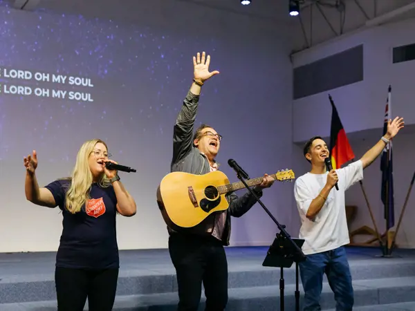 Three people sing with spirit, accompanied by one person playing the guitar.