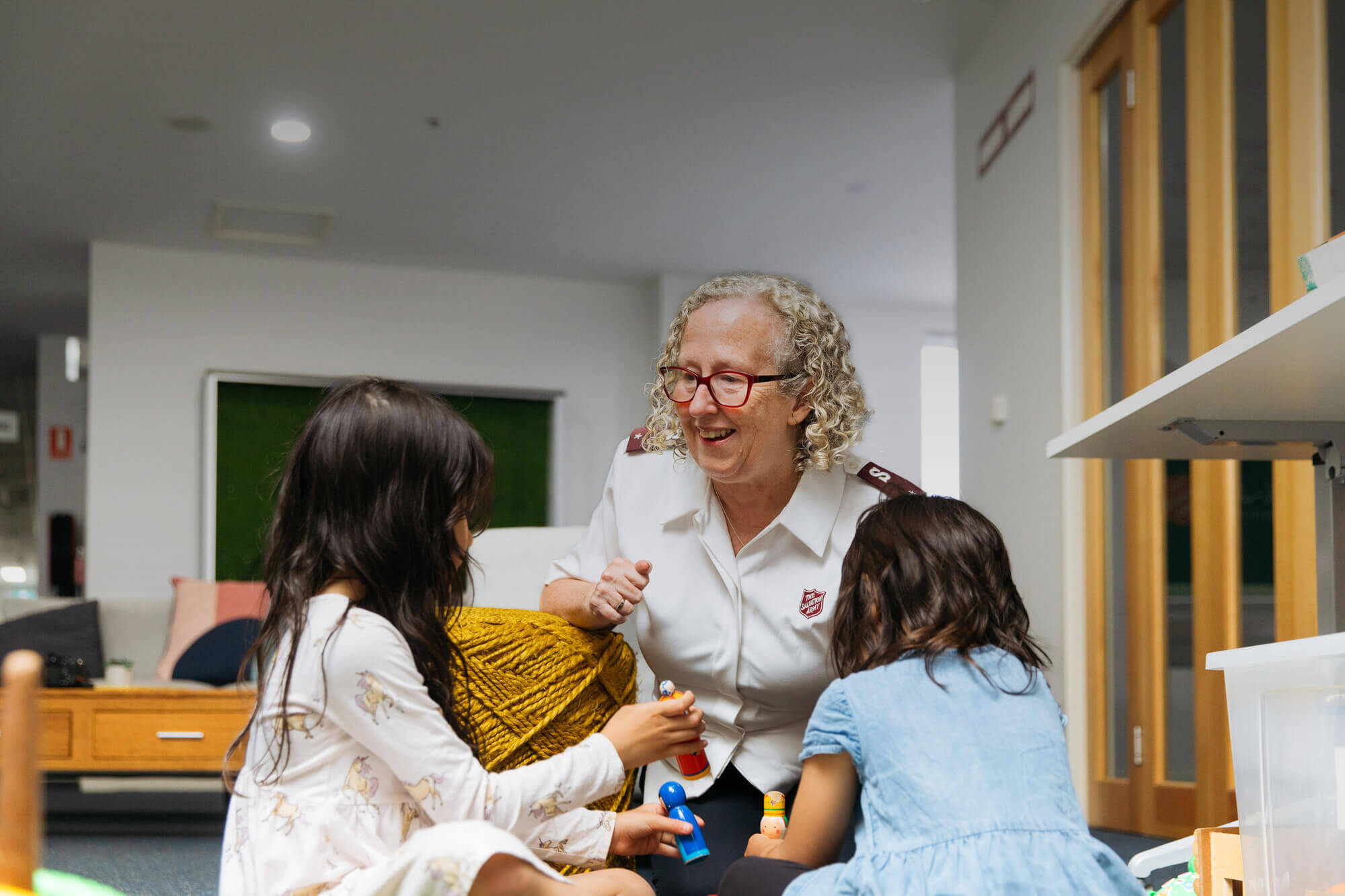 Two children playing with toys as female Salvos Officer is smiling.