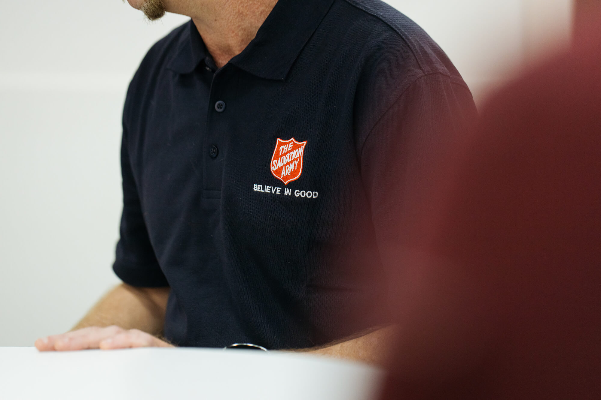 A smiling person wearing a Salvation Army polo shirt sitting at a welcome table.