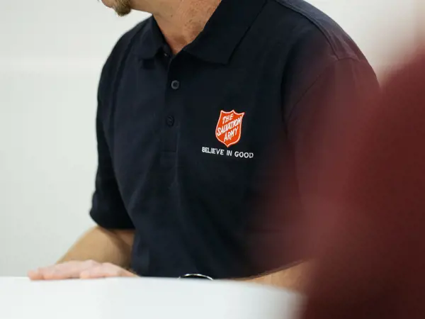 A smiling person wearing a Salvation Army polo shirt sitting at a welcome table.