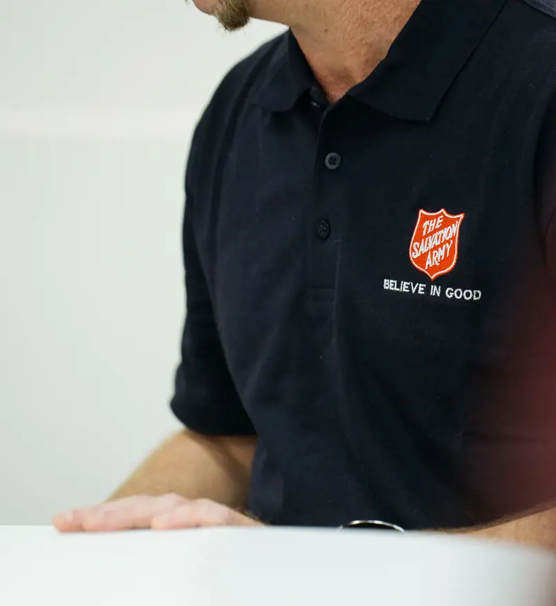 A smiling person wearing a Salvation Army polo shirt sitting at a welcome table.