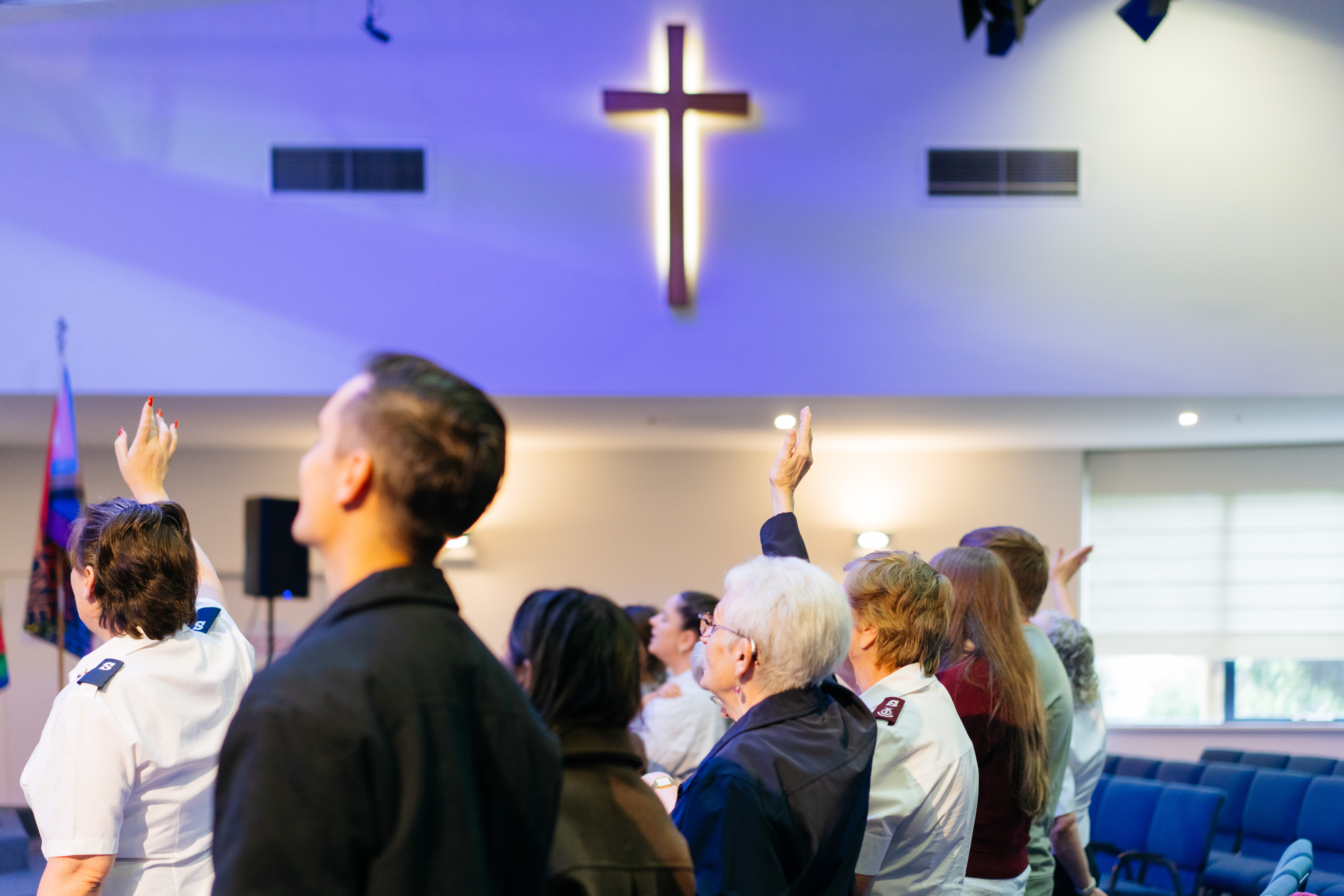 Several people, including a man, a woman with gray hair, and a young woman, are raising their hands in a church. A large, a cross is illuminated on the ceiling. Salvation Army flags are visible in the background.