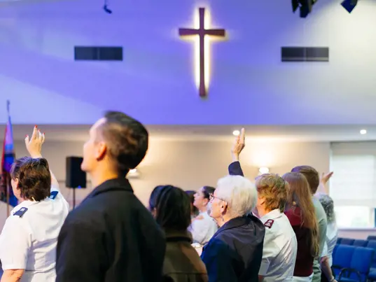 Several people, including a man, a woman with gray hair, and a young woman, are raising their hands in a church. A large, a cross is illuminated on the ceiling. Salvation Army flags are visible in the background.