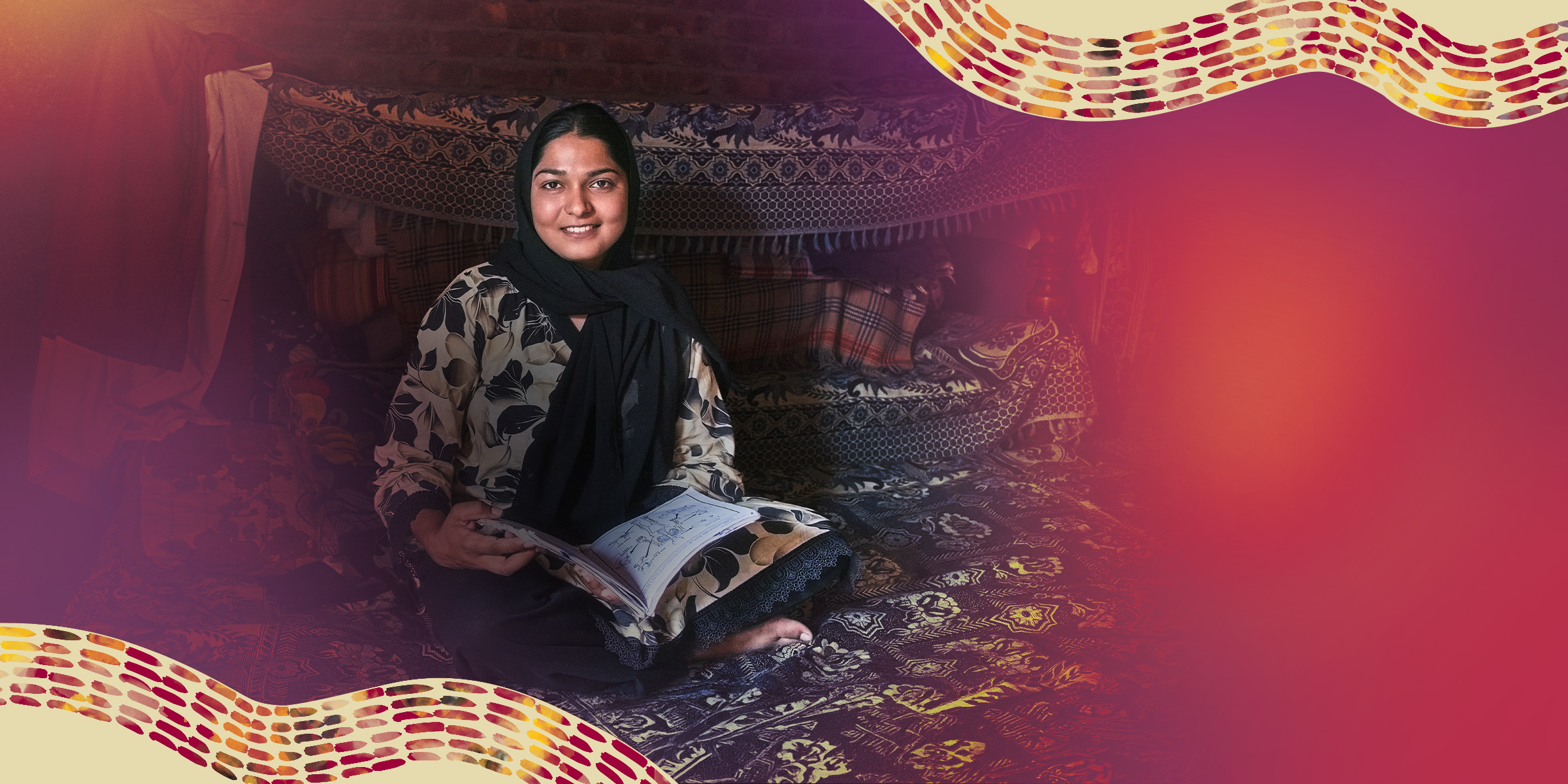 Woman smiling seated on patterned rugs inside a fabric-lined shelter, holding an open notebook and looking toward the camera.