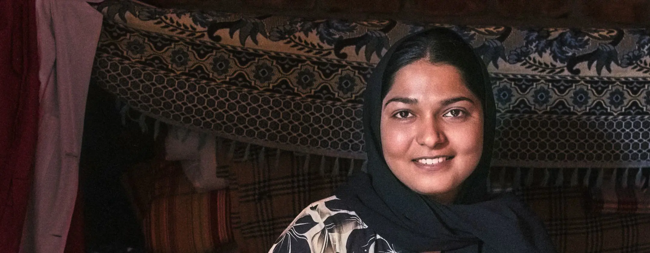 A woman smiles as sits on a bed surrounded by beautifully-woven Pakistani textiles.