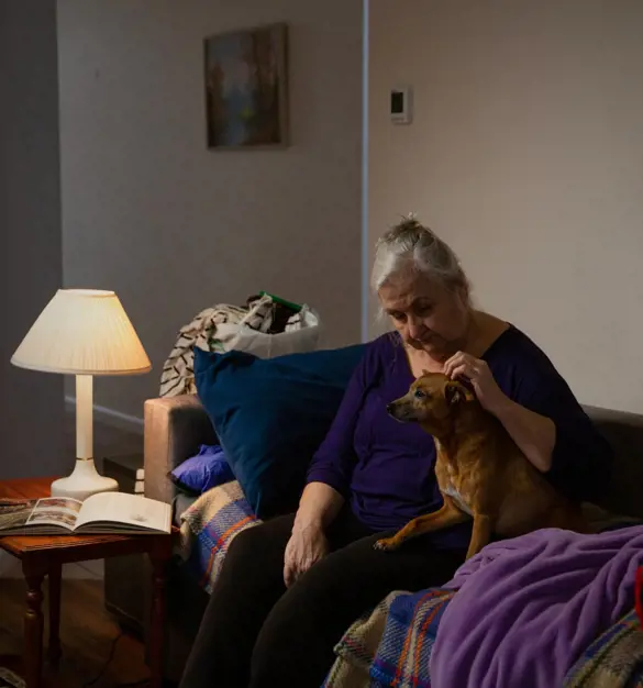 Elderly person on a couch petting a brown dog in a softly lit living room.