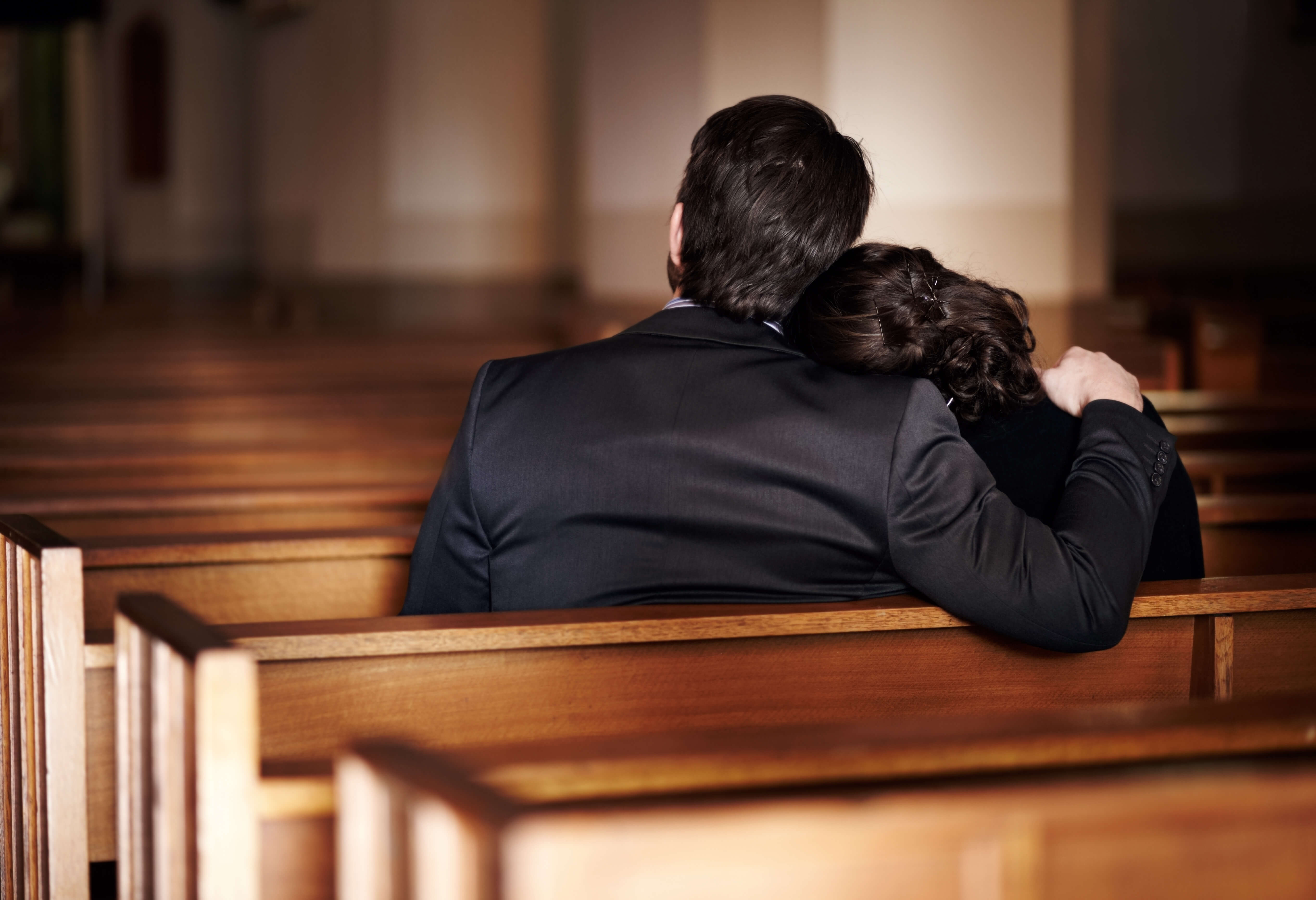 Two people sit together on a wooden church pew, viewed from behind, with one person’s arm around the other in a softly lit interior.